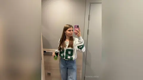 Young woman in a grey graphic sweatshirt and blue jeans taking a mirror selfie in a modern minimalist hallway.