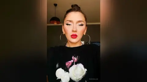 Woman with bold red lipstick and winged eyeliner wearing a black floral graphic t-shirt in a dimly lit room.