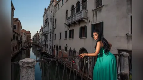 MissyJolie poses on a stone balcony overlooking a Venice canal, wearing a long teal dress that accentuates her curves.