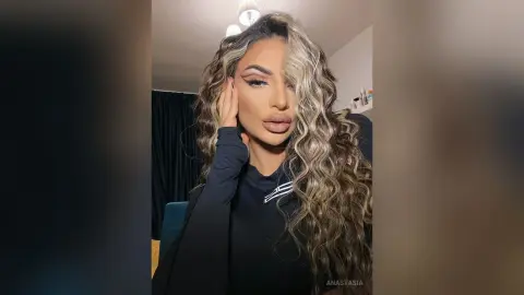 Anastasia poses with a hand to her face, wearing a black long-sleeved top and showing off her long, wavy blonde and brown hair.