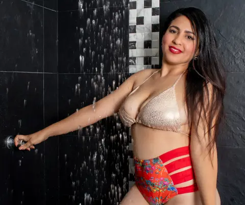 Smiling woman in a gold bikini and red patterned swimsuit standing under a shower in a dark tiled bathroom.