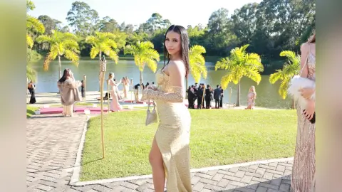 Elegant woman in a gold sequin evening gown posing outdoors by a lake at a sunny garden party.
