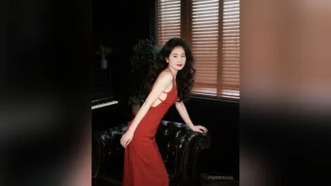 Elegant woman in a red evening gown posing in a moody, dark interior by a window with blinds.