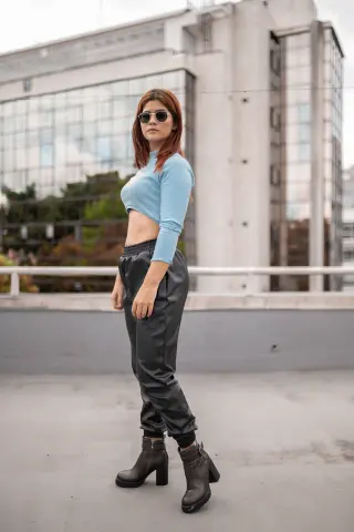 Stylish woman in a light blue crop top and black leather trousers posing on an urban rooftop.