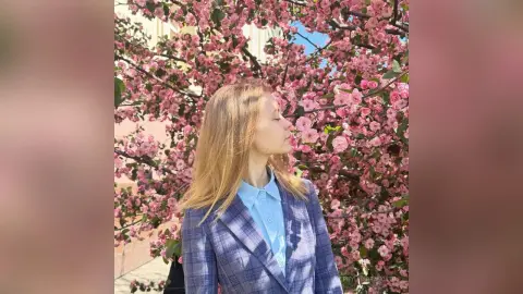Blonde woman in a blue plaid blazer smiling amidst blooming pink cherry blossom trees in a dreamy spring setting.