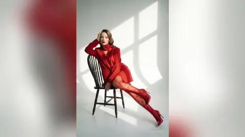 Elegant woman in a red blazer and patterned tights sitting on a chair in bright studio lighting.