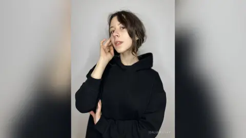 Young woman in a black hoodie posing against a plain white background with a relaxed, casual mood.