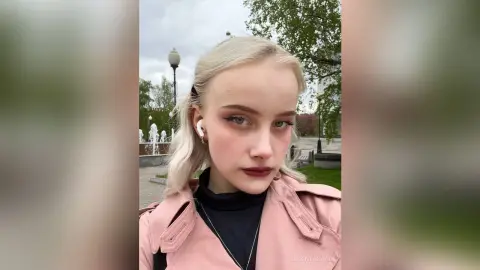 Blonde woman in a pink trench coat and black turtleneck posing outdoors near a park fountain.
