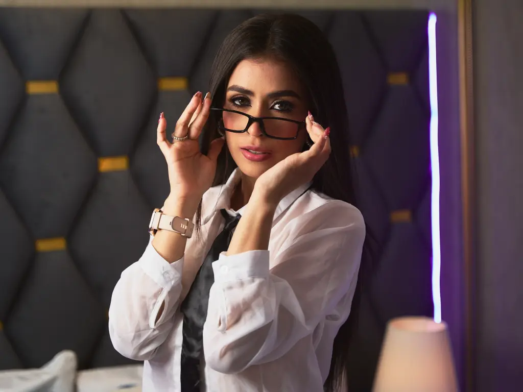 VictoriaKorz poses wearing glasses and a white button-down shirt, running her hands up her face with long dark hair.