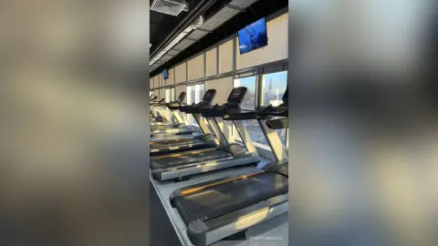 ChristineDavis poses in a gym setting lined with several treadmills next to large windows overlooking a city view.
