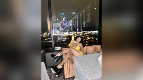 NicoleBella sits on a peach-colored sofa wearing a yellow lace bodysuit and black platform heels against a night city view.