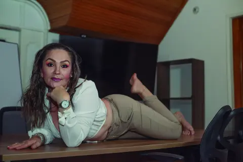 Smiling woman in a white blouse and beige trousers lying on a wooden table in a modern indoor setting.