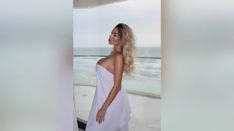 Blonde woman in a white strapless dress posing on a luxury balcony overlooking the ocean at sunset.