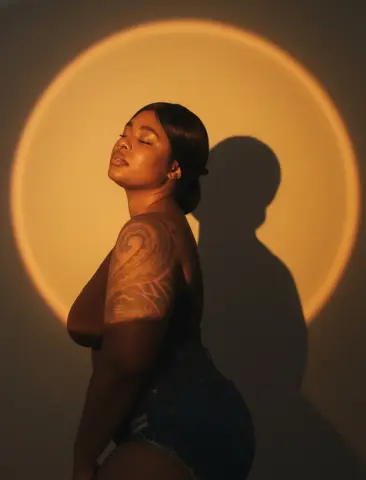 Curvy woman with tattoos posing in denim shorts against a warm golden circular light shadow.
