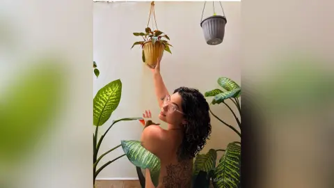 Smiling woman with curly hair in a minimalist white room surrounded by lush green indoor plants.