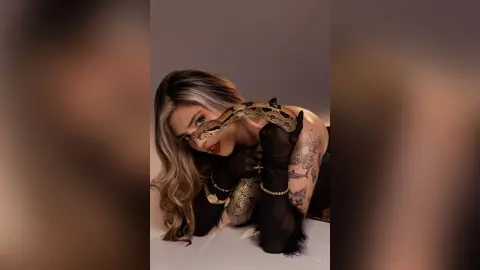 Glamorous woman in a black mesh outfit holding a large snake against a neutral studio background.
