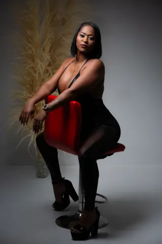 Elegant woman in a black bodysuit and high heels sitting on a red chair against a soft pampas grass background.