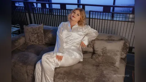 Blonde woman in white silk pajamas relaxing on a plush sofa on a luxury balcony at twilight.