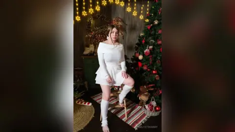 Young woman in a white festive outfit posing by a decorated Christmas tree in a cozy holiday setting.