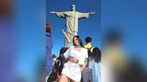 Alison poses in a white crop top and skirt with her arm raised against a large stone statue under a clear blue sky.