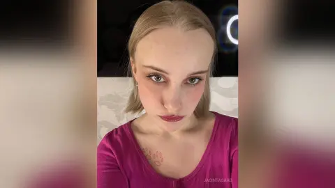 Blonde woman in a pink v-neck top with soft makeup posing in a dimly lit indoor setting.