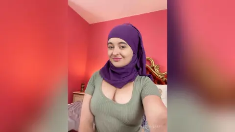 Smiling woman in a purple hijab and sage green ribbed top posing in a pink bedroom.