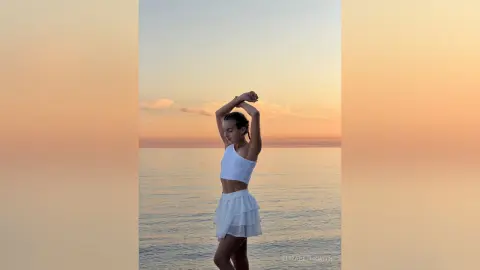 Young woman in a white summer outfit standing by the ocean during a peaceful golden hour sunset.