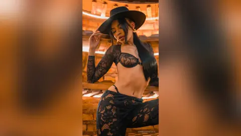 Stylish woman in black lace lingerie and a wide-brimmed hat posing in warm, moody indoor lighting.