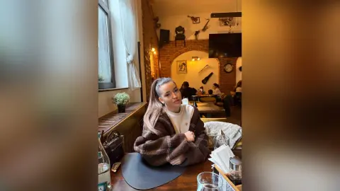 Young woman in a cozy brown sweater sitting in a warm, rustic cafe setting with a relaxed mood.