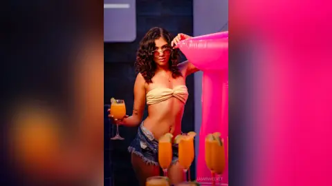 Smiling woman in a yellow bandeau top and denim shorts holding a cocktail at a vibrant pink summer party.