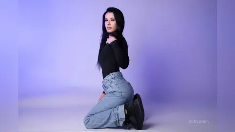 Young woman in a black long-sleeve top and light blue jeans posing against a purple studio background.