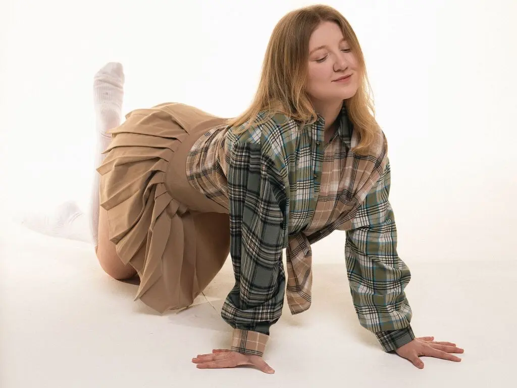 Smiling young woman in a plaid shirt and tan pleated skirt posing against a white background.