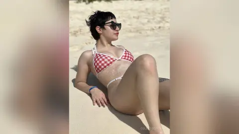 Woman in red and white gingham bikini relaxing on a sunny beach wearing sunglasses.