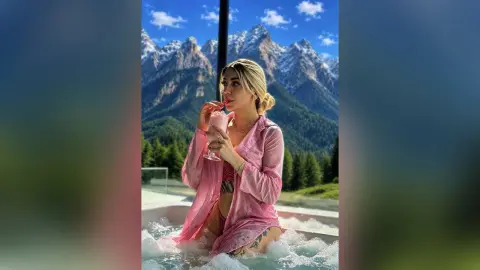 Blonde woman in a pink robe sipping a drink in a luxury outdoor hot tub with mountain views.