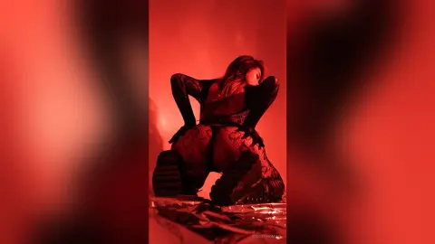 RosieThomas — RosieThomas poses in a black lace bodysuit and black boots, kneeling against a red background with her hair falling over her face.