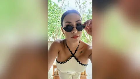 Smiling woman in a white scalloped halter top and sunglasses outdoors in a sunny garden setting.