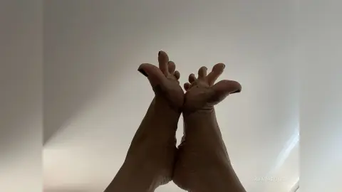 Close-up of bare feet with dark nail polish held up against a plain white background in a minimalist setting.