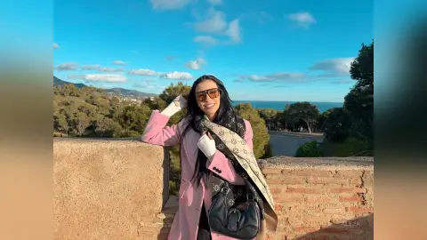 Aleja smiles while wearing a pink coat and a patterned scarf, standing outdoors against a view of trees and the ocean.