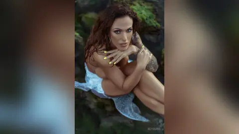 ReyQuins poses outdoors in a white lace outfit, showing off her long brown hair and tattoos while sitting in a lush green setting.