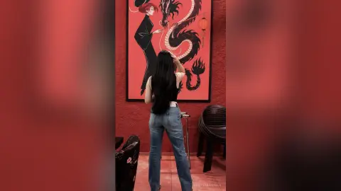 Woman in blue jeans standing before a large dragon painting in a red room.