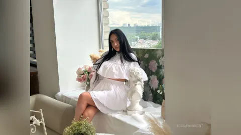 TessaWinsent — TessaWinsent sits on a white bed by a window wearing a white off-the-shoulder crop top and skirt.