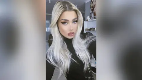 Blonde woman with winged eyeliner wearing a black turtleneck in a bright indoor setting.