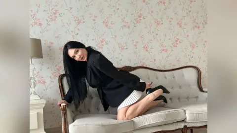 TessaWinsent — TessaWinsent in a black blazer and white striped skirt poses on a vintage white sofa against floral wallpaper.