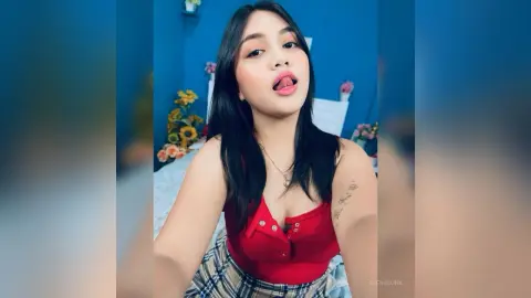 Young woman in a red tank top and plaid skirt posing playfully against a blue wall background.
