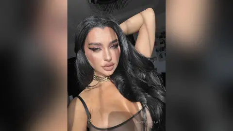 NicoleBella poses with her hand behind her head, wearing a black bra and a gold choker necklace.