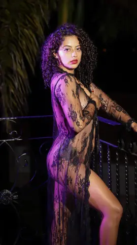 Curvy woman in a sheer black lace robe posing outdoors at night with tropical palm leaves.
