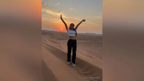 Joyful woman in white crop top and black pants celebrating at sunset in a vast desert landscape.
