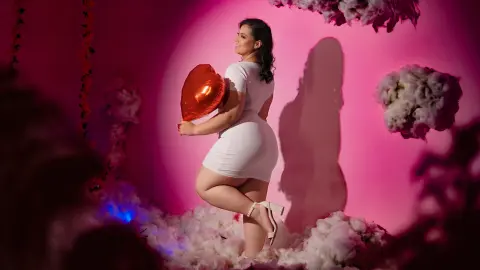 Smiling woman in a white mini dress holding a red heart balloon against a pink floral studio backdrop.