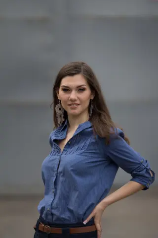 Smiling woman in a blue button-down shirt and large silver earrings standing against a neutral gray background.