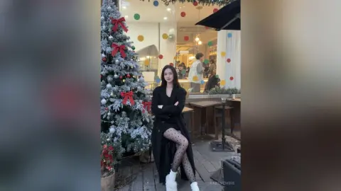 Stylish woman in a black coat and polka dot tights posing by a decorated Christmas tree in a cozy cafe setting.